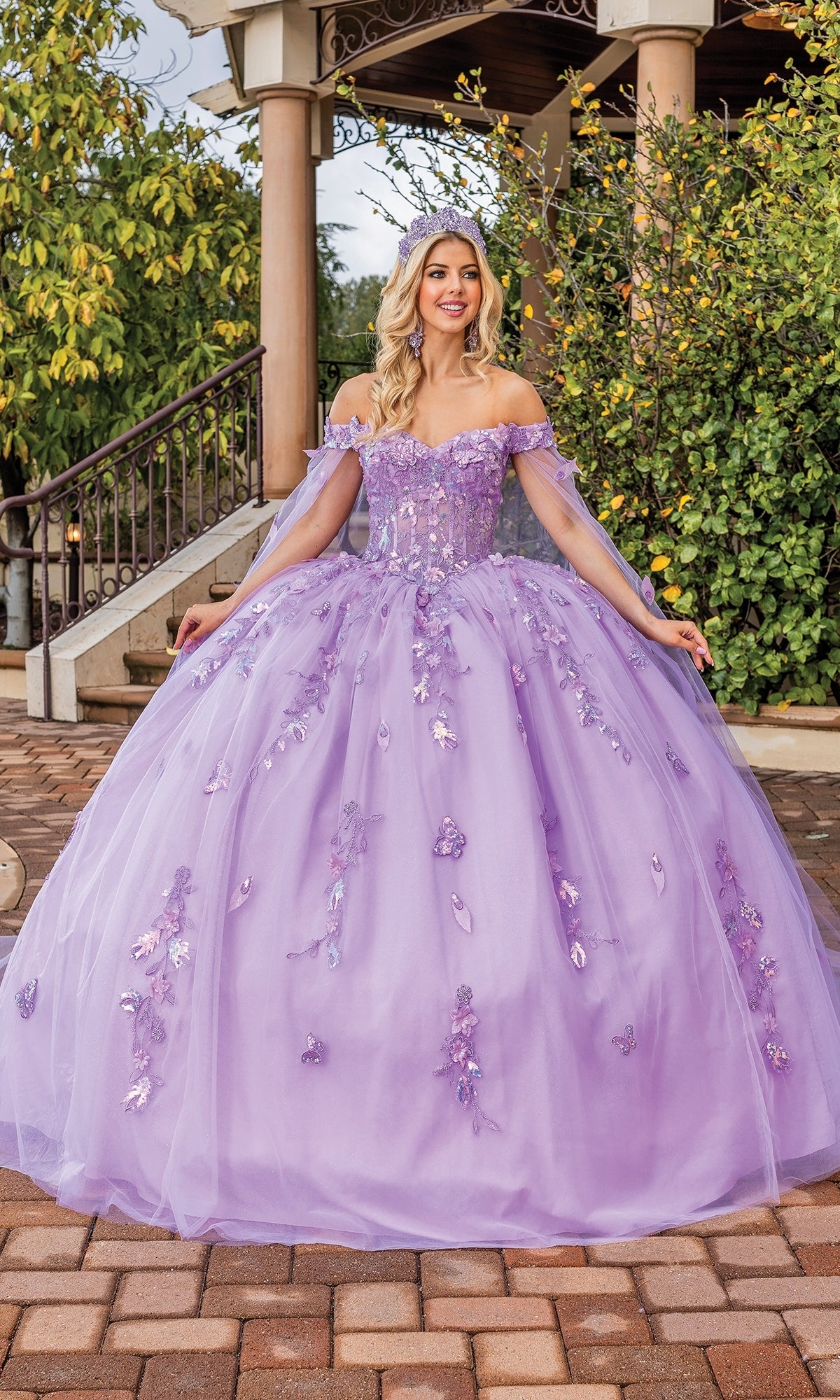 A woman stands outdoors smiling in front of greenery and a stone staircase, wearing the Quinceanera Dress 1840 By Dancing Queen—an off-the-shoulder lavender ball gown with floral embellishments and a sweetheart neckline.